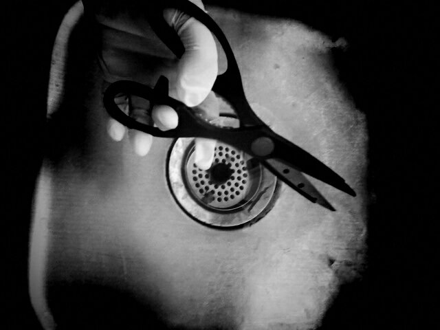 Black and white photoof gloved hand holding scissors over a sink.