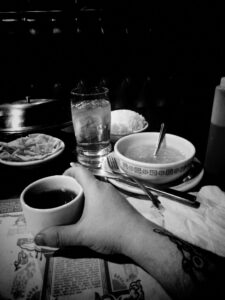 Black and white photo of table with food dishes on it. Arm reaching for cup.