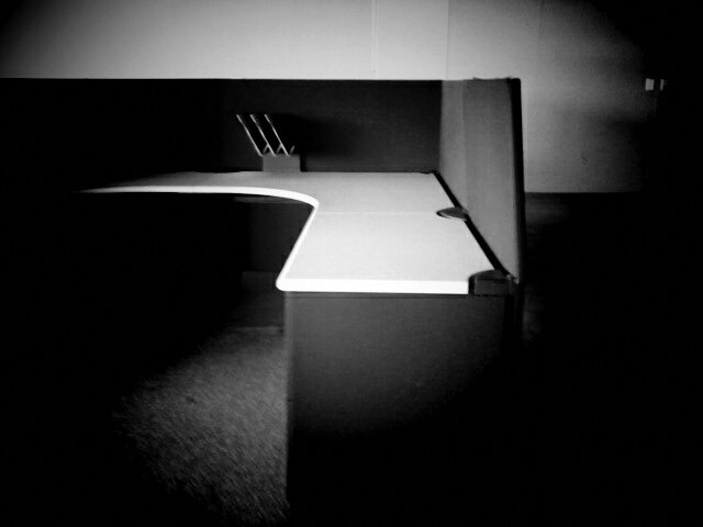 Black and white photo of desk in low walled cubicle.