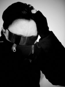 Black and white photo/self portrait wearing shades. One hand is holding my head. I am wearing a hoodie that has a cowl obscuring half my face.