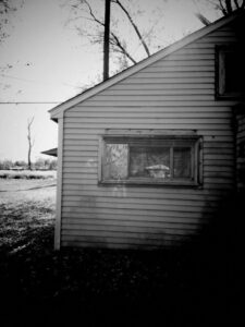 Black and white photo of left side of dilapidated dirty house. Train tracks and trees are behind the house.