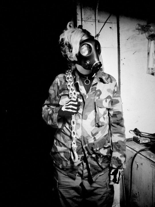 Black and white photo of a person wearing a camouflage shirt, leather collar with ring, cargo pants, skeleton gloves, holding a thick heavy chain over one shoulder. Gas mask covering face with blonde hair draped over mask. They are standing in a garage.