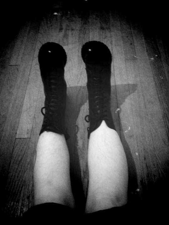 Black and white photo of legs on dirty wood floor. Shown from the knees down to the feet of the leather combat jump boots.