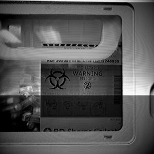 Black and white photo of sharps container with biohazard warning. Photo has a glitched effect with lines through it.