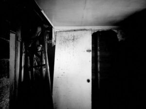 Black and white photo of decaying building from the inside. The walls are dark, the ceiling is a dirty white and the door is white and off the hinges.