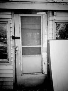 Black and white photo of locked screen door on dilapidated building.