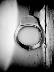 Black and white photo of handcuffs hanging against paint peeling inside a closet.