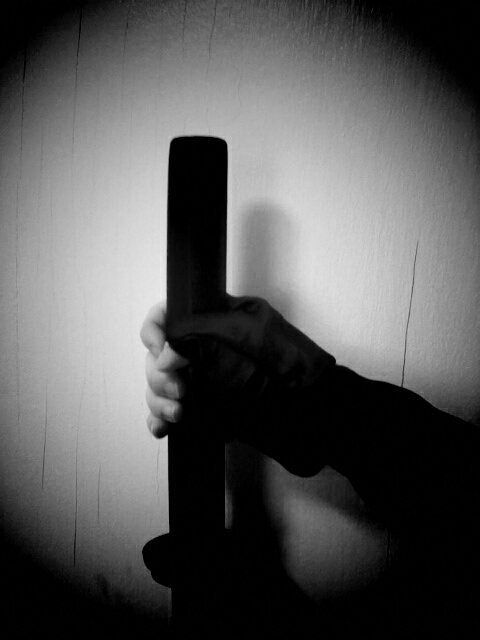 Black and white photo of hand wearing fingerless glove holding on to a sword. Background is a white cracked wall.