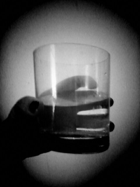 Black and white photo of hand with chipped black nail-polish holding an unusual shaped glass filled halfway with a fluid.