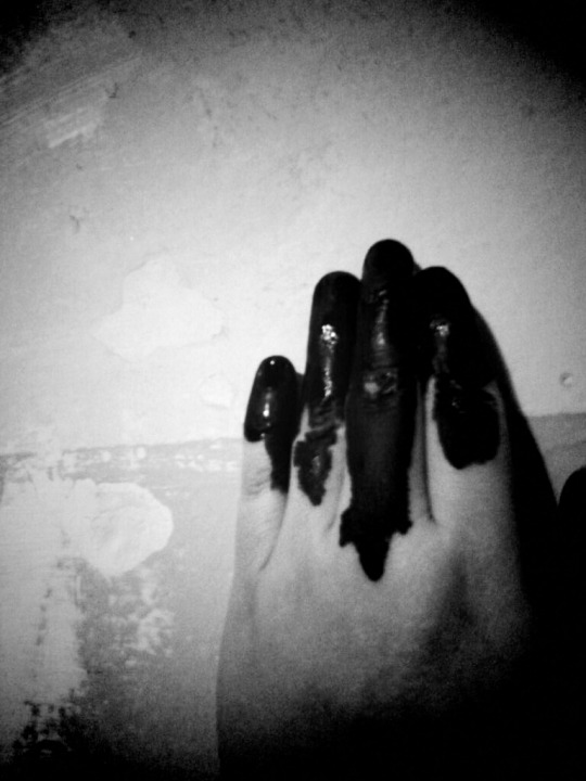 Photo of hand covered with black glistening paint against a peeling wall.