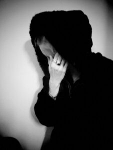 Black and white photo of person hiding their face with their hand. They are wearing a black hooded jacket.