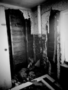 Black and white photo of inside of destroyed home. Broken walls with wood slats exposed.