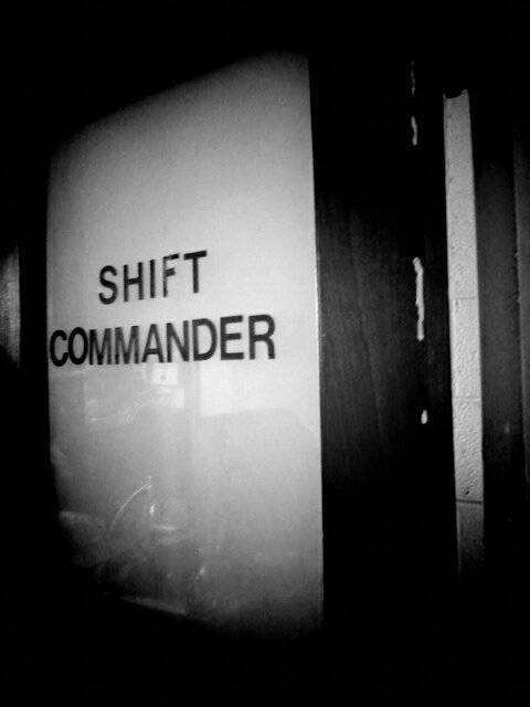 Black and white photo of door. Glass on door has the words shift commander.