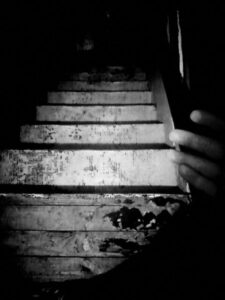 Black and white photo of stairs leading down with hand. Stairs are stripped, dirty and have paint marks on them.