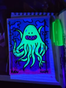 Photo of marker art on a sticker depicting a happy tentacle type creature with an open smiling mouth and a few teeth under UV black light.