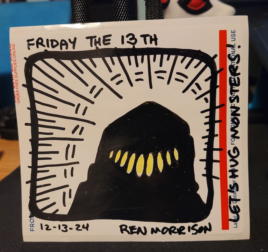 Upclose photo of drawing marker art on a sticker depicting a a shadowy figure with teeth. Title "Let's Hug Monsters!" Drawn on Friday the 13th.